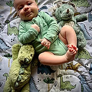 Glenn joined the competition — help win amazing prizes! baby, infant, onesie, green_clothing, plush_toy, dinosaur_plush, blanket, dinosaur_pattern, cute, drool, smiling, lying_down, child, soft_toy, toddler, indoor, playtime, happy, face, skin