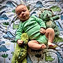 Glenn is registered to the contest to win money with this photo: baby, infant, dinosaur, plush_toy, blanket, onesie, cute, child, soft_toy, green_clothing, lying_down, indoor, portrait, toddler, baby_toy, playtime, smiling, cute_expression, patterned_blanket, hand_grip