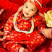 Brijesh joined the competition — help win amazing prizes! baby, child, traditional_clothing, red_outfit, hat, pearl_necklace, balloon, hands, fabric, sitting, portrait, cute, celebration, festive, infant, smiling, indoors, person, cultural, bright_colors