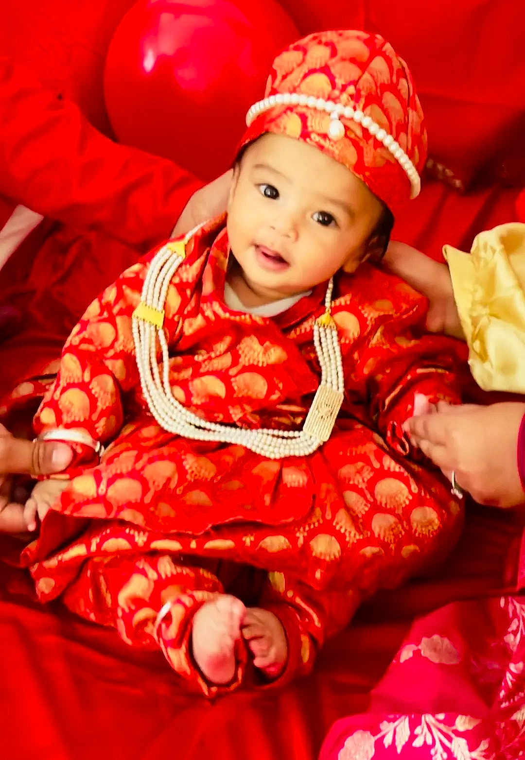 Brijesh joined the competition — help win amazing prizes! baby, child, traditional_clothing, red_outfit, hat, pearl_necklace, balloon, hands, fabric, sitting, portrait, cute, celebration, festive, infant, smiling, indoors, person, cultural, bright_colors