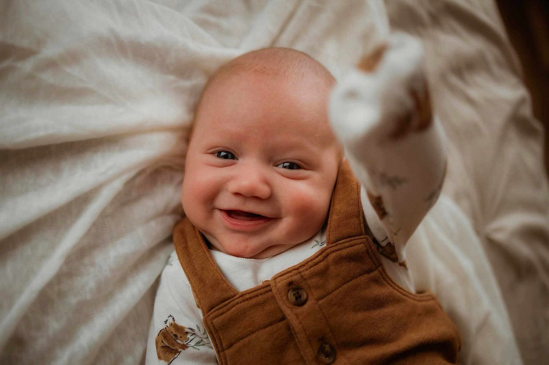Waylon joined the competition — help win amazing prizes! baby, infant, smiling, face, cheeks, close_up, onesie, overalls, brown_overalls, blanket, bedding, soft_light, portrait, newborn, cute, small_hand, sock, fabric, cozy, indoor