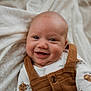 baby, infant, smiling, face, eyes, blanket, bedding, clothing, overalls, onesie, portrait, close_up, happy, adorable, nursery, skin, head, bed, cozy, candid