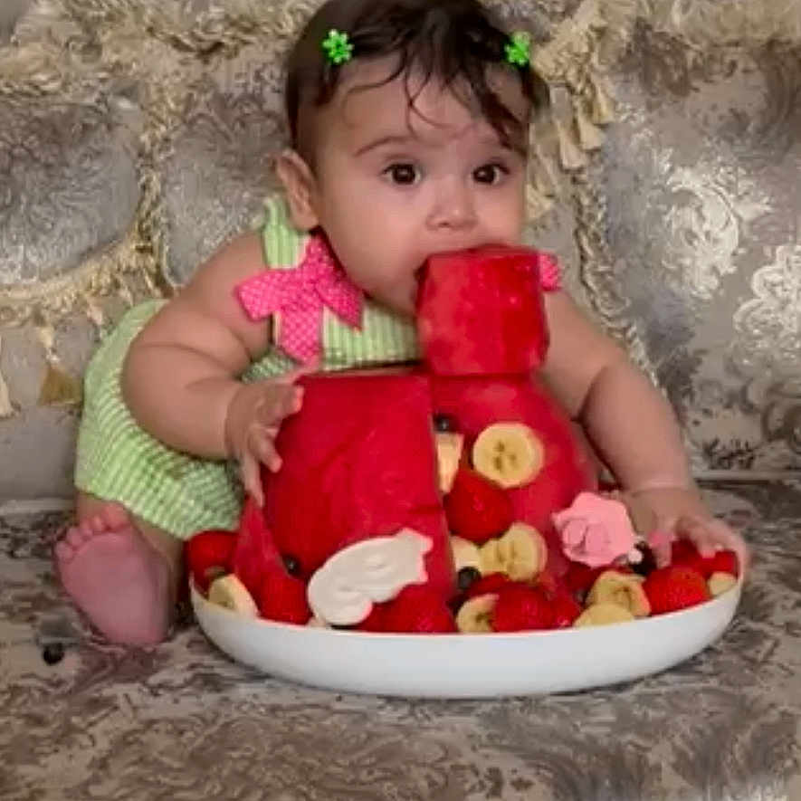 Nazli joined the competition — help win amazing prizes! baby, banana, child, colorful, couch, curious, cute, food, fruit, green_dress, hair_clips, indoor, patterned_furniture, playful, portrait, sitting, snack, strawberries, summer, watermelon