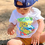 Ezio participe au concours pour gagner de l'argent avec cette photo : toddler, child, cap, blue_cap, white_tshirt, colorful_tshirt, sitting, gravel, outdoor, sunlight, shorts, yellow_shorts, shoes, white_shoes, hands, face, eyelashes, focused, casual_clothing, summer