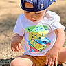toddler, child, cap, blue_cap, white_tshirt, colorful_tshirt, sitting, gravel, outdoor, sunlight, shorts, yellow_shorts, shoes, white_shoes, hands, face, eyelashes, focused, casual_clothing, summer