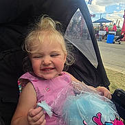 Miley is registered to the contest to win money with this photo: toddler, child, stroller, cotton_candy, pink_dress, smiling, outdoor, fair, blonde_hair, happy, plastic_bag, people, flags, sky, clouds, grass, event, fun, daytime, casual