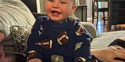 Spencer is registered to the contest to win money with this photo: baby, infant, smile, child, onesie, football_pattern, couch, living_room, adult, hand, table, chair, vase, flowers, curtains, lamp, door, carpet, person, home_interior