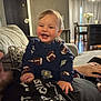 baby, infant, smile, child, onesie, football_pattern, couch, living_room, adult, hand, table, chair, vase, flowers, curtains, lamp, door, carpet, person, home_interior