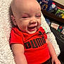 baby, infant, smiling, red_shirt, puma_logo, blanket, happy, face, child, indoors, furniture, bookshelf, books, clothing, person, portrait, sitting, cute, young_child, cozy