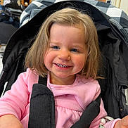 Kathaléna a rejoint le concours — aidez-le/la à gagner de superbes lots ! toddler, child, smile, stroller, pink_clothing, bracelet, indoor, person, happy, face, blue_eyes, hair, seatbelt, baby, cute, cozy, hand, plant, background, accessory