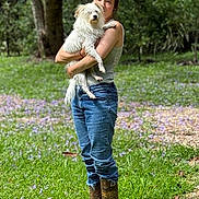 Rosie is registered to the contest to win money with this photo: person, dog, grass, boots, jeans, tank_top, outdoor, tree, flowers, nature, pet, animal, holding, casual, portrait, daylight, greenery, fur, cute, companion