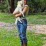 person, dog, grass, boots, jeans, tank_top, outdoor, tree, flowers, nature, pet, animal, holding, casual, portrait, daylight, greenery, fur, cute, companion