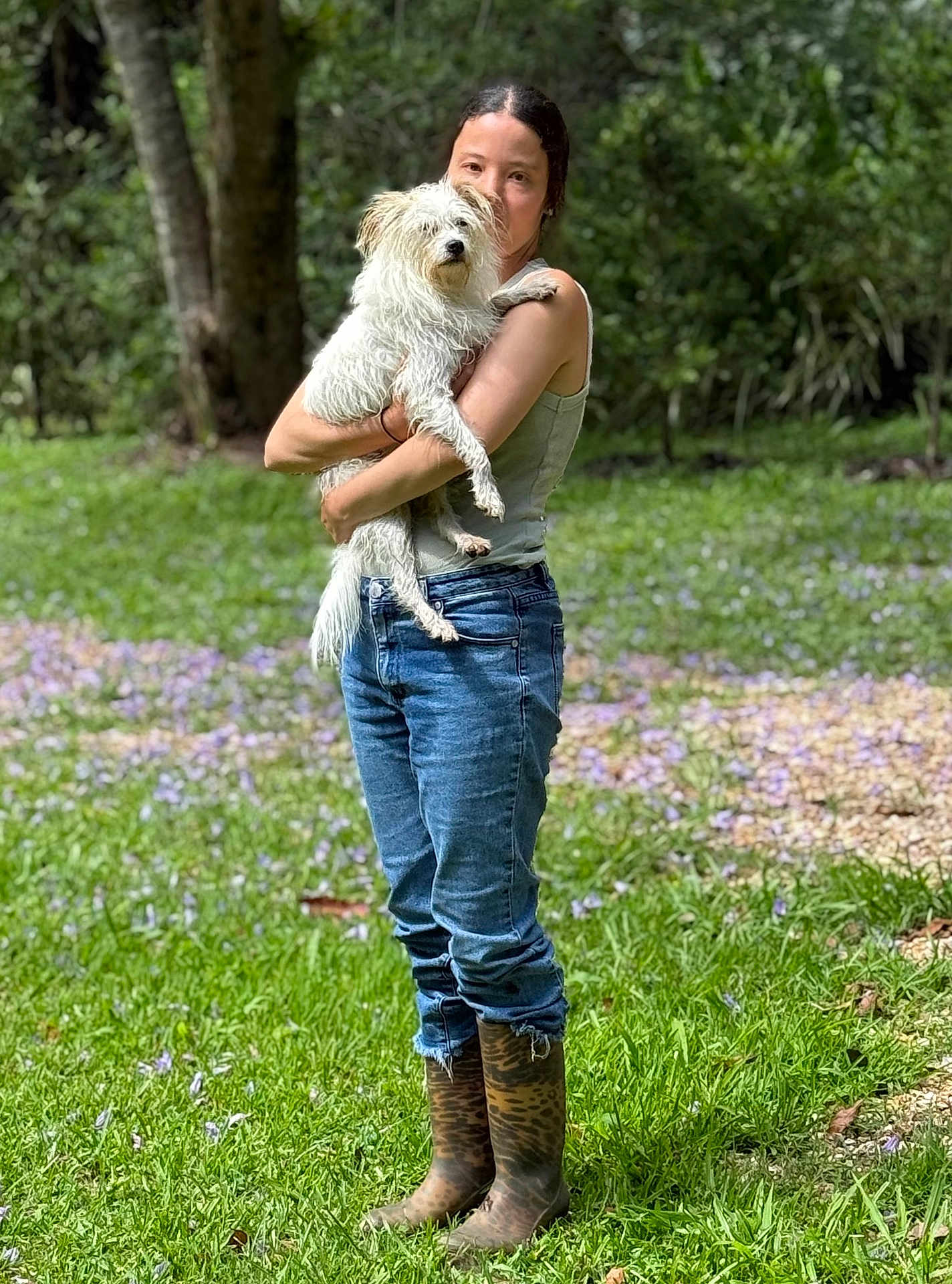 Rosie is registered to the contest to win money with this photo: person, dog, grass, boots, jeans, tank_top, outdoor, tree, flowers, nature, pet, animal, holding, casual, portrait, daylight, greenery, fur, cute, companion