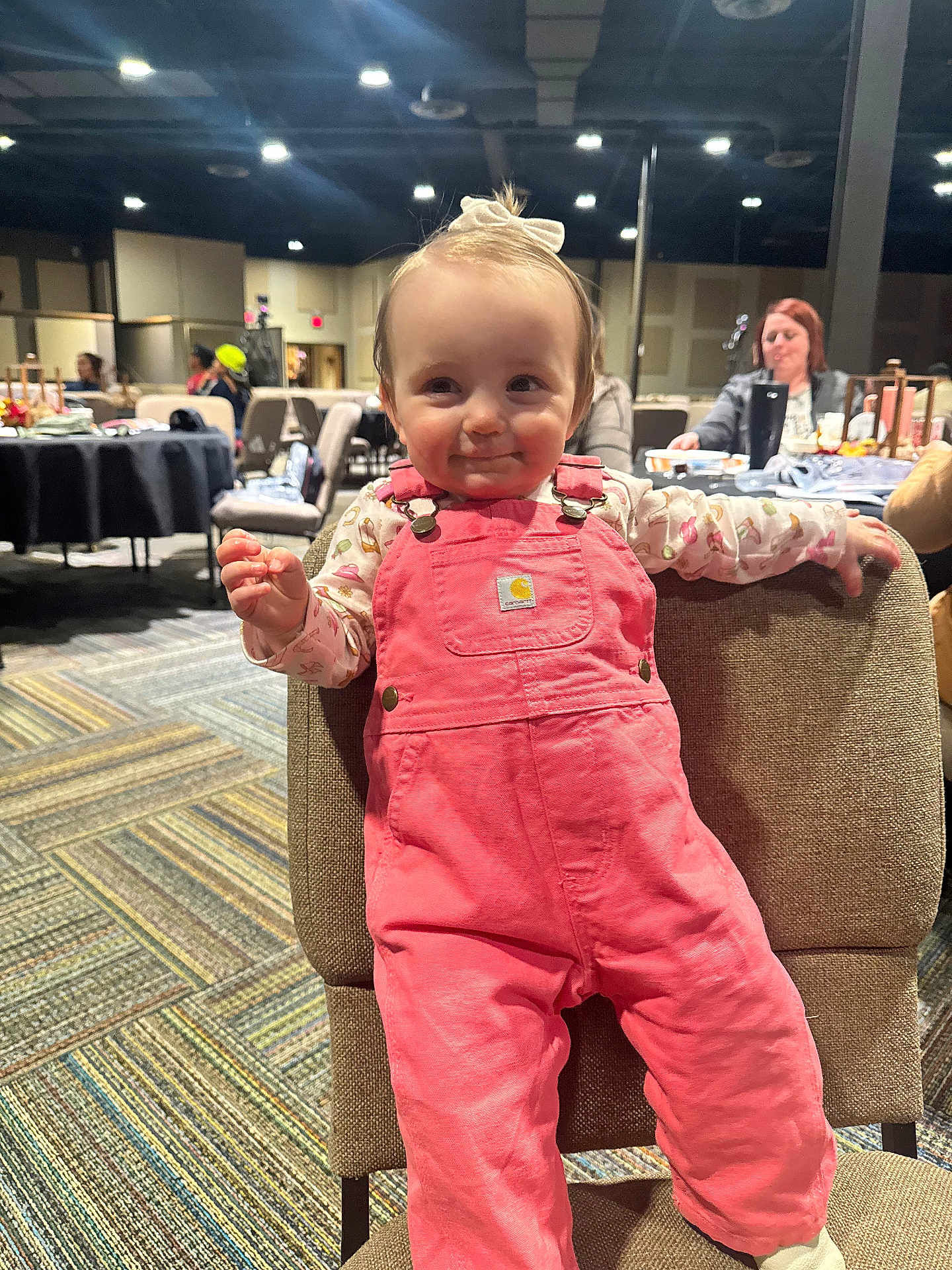 Elowyn joined the competition — help win amazing prizes! baby, child, pink_overalls, smile, indoor, chair, carpet, people, woman, background, lighting, face, person, clothing, cute, happy, seated, headband, toddler, room