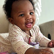 Asmara joined the competition — help win amazing prizes! baby, child, infant, smiling, happy, curly_hair, onesie, heart_pattern, indoor, soft_surface, close_up, cute, playful, person, skin, hand, face, tongue_out, couch, pacifier