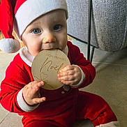 Gauthier participe au concours pour gagner de l'argent avec cette photo : baby, child, santa_hat, red_clothing, blue_eyes, indoor, floor, wooden_plaque, portrait, holiday, christmas, cute, smiling, sitting, hand, face, plush, cozy, person, festive