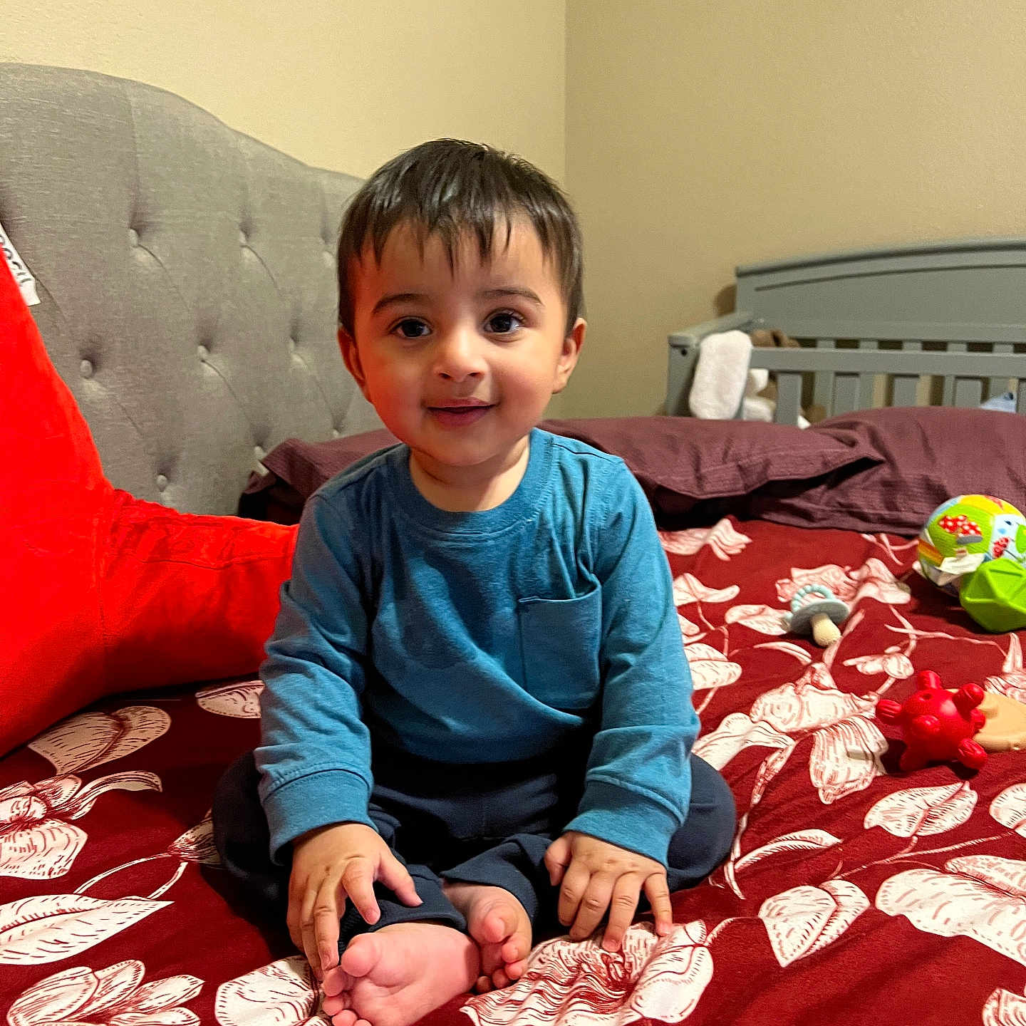 Arnaaz joined the competition — help win amazing prizes! bed, blue_shirt, child, cozy, crib, face, foot, furniture, hand, happy, headboard, home, indoor, patterned_bedspread, person, pillow, sitting, smile, toddler, toy