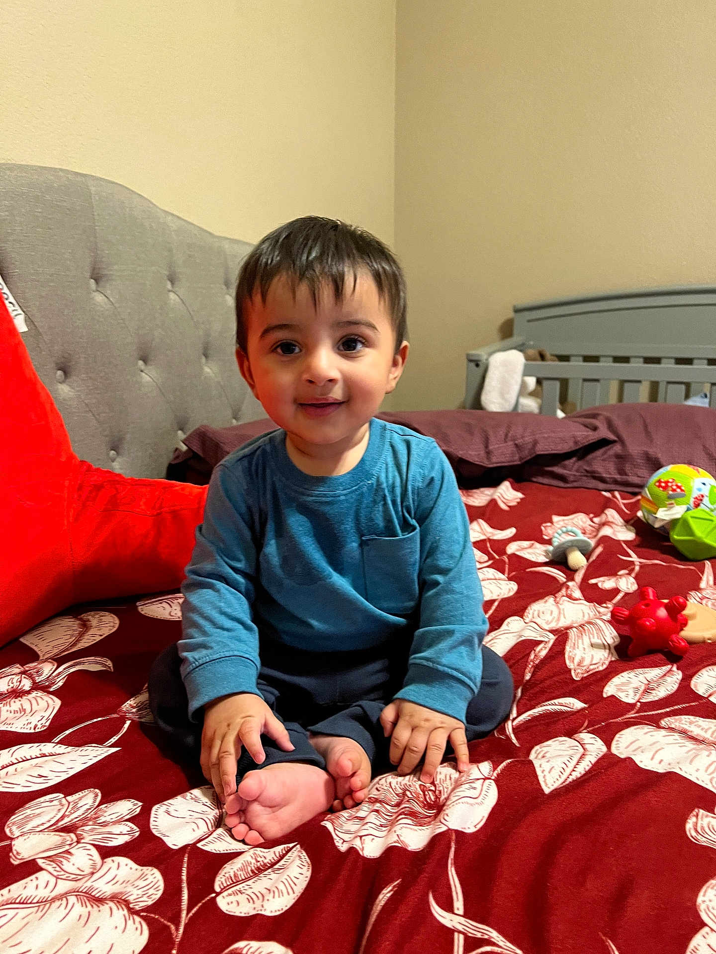 Arnaaz joined the competition — help win amazing prizes! toddler, child, bed, toy, blue_shirt, smile, indoor, furniture, crib, headboard, pillow, foot, hand, face, person, home, cozy, happy, sitting, patterned_bedspread