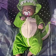Chloe is registered to the contest to win money with this photo: baby, bare_feet, bed, blanket, child, costume, cozy, cute, dinosaur_costume, dragon, face, green_clothing, hood, indoor, infant, lying_down, pacifier, patterned_bedding, portrait, sleeping_area