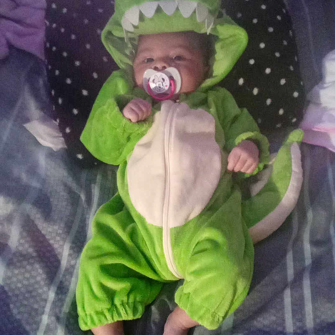 Chloe is registered to the contest to win money with this photo: baby, bare_feet, bed, blanket, child, costume, cozy, cute, dinosaur_costume, dragon, face, green_clothing, hood, indoor, infant, lying_down, pacifier, patterned_bedding, portrait, sleeping_area