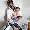 man, child, toddler, indoor, casual_clothing, sunglasses, cap, smile, seated, carpet, white_hoodie, colorful_text, yellow_shirt, jeans, watch, ring, hand_holding, table, chair, wall