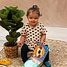 toddler, child, book, toy, plant, potted_plant, rug, floor, chair, indoors, cute, smiling, clothing, hair_tie, patterned_rug, colorful, illustration, sitting, playtime, background