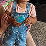 Araya is registered to the contest to win money with this photo: adult_hand, barefoot, blanket, carpet, child, couch, curly_hair, denim_overalls, footwear, furniture, happy, indoor, living_room, person, short_sleeve_shirt, smiling, sock, television, toddler, toy