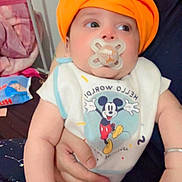 Jorawar is registered to the contest to win money with this photo: baby, pacifier, orange_turban, bib, mickey_mouse, person, hand, bed, blanket, clothing, infant, cute, indoor, holding, face, expression, young_child, arm, sitting, comfort