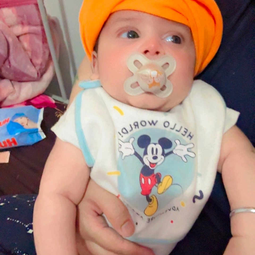 Jorawar is registered to the contest to win money with this photo: arm, baby, bed, bib, blanket, clothing, comfort, cute, expression, face, hand, holding, indoor, infant, mickey_mouse, orange_turban, pacifier, person, sitting, young_child
