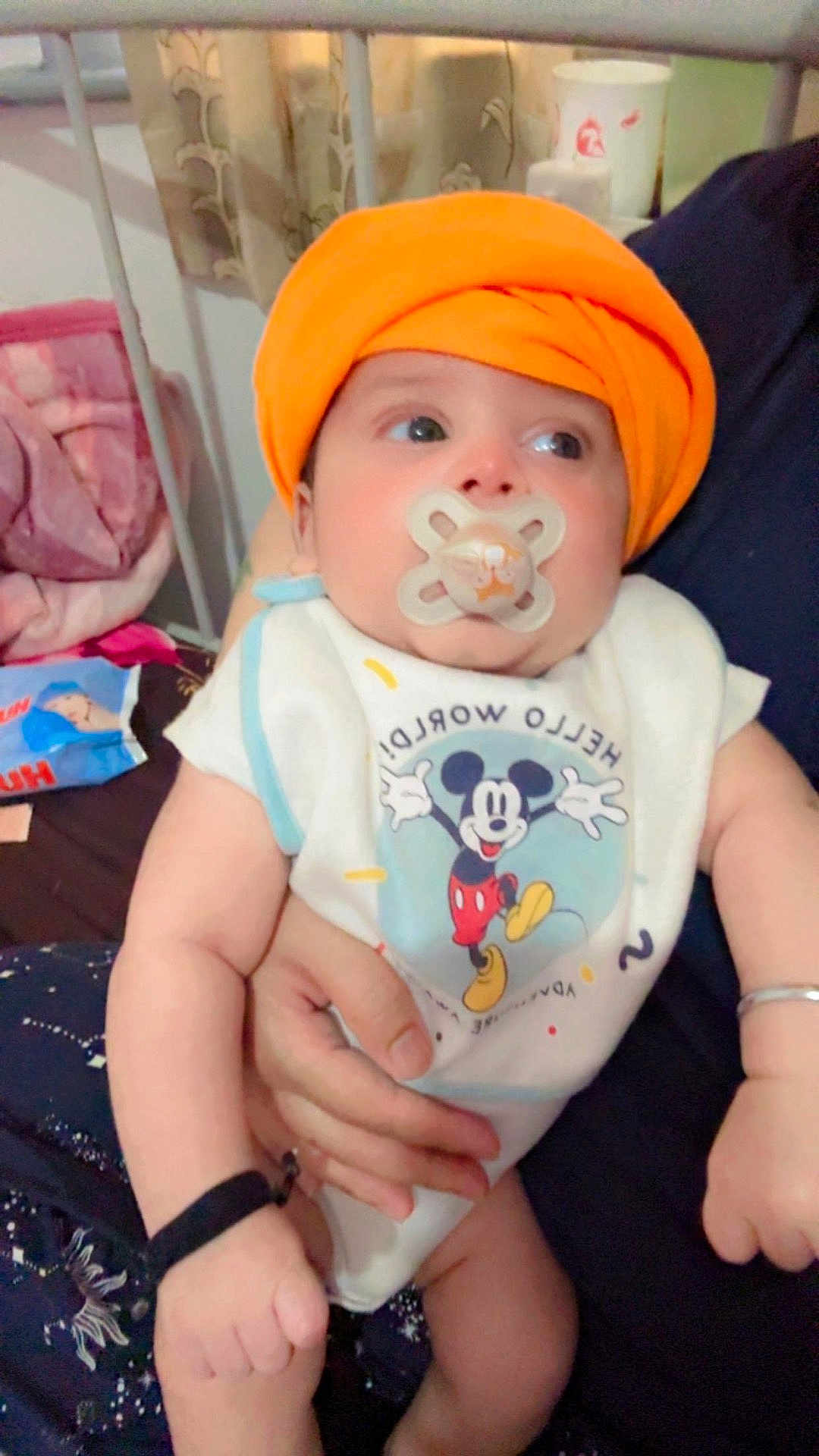 Jorawar is registered to the contest to win money with this photo: baby, pacifier, orange_turban, bib, mickey_mouse, person, hand, bed, blanket, clothing, infant, cute, indoor, holding, face, expression, young_child, arm, sitting, comfort