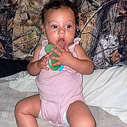 Journey is registered to the contest to win money with this photo: baby, child, infant, teether, pink_onesie, sitting, bed, pillow, woodland_pattern, curly_hair, indoor, person, cute, chewing, skin, face, expression, young, furniture, blanket
