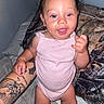 baby, child, infant, smiling, pink_onesie, tattooed_arm, hand, bed, pillow, camo_pattern, indoor, person, cute, joyful, skin, face, happy, standing, support, expression