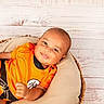 baby, infant, smile, orange_clothing, basket, cushion, wooden_background, portrait, happy, child, cute, relaxed, indoors, person, head, face, sitting, bright, soft_lighting, cozy