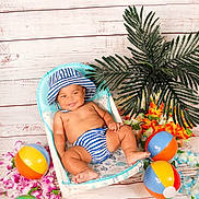 William is registered to the contest to win money with this photo: baby, child, striped_hat, striped_diaper, beach_balls, tropical_flowers, palm_tree, chair, smiling, happy, indoor, decor, playful, summer_theme, cute, infant, colorful, portrait, relaxing, fun