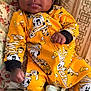 Aurora is registered to the contest to win money with this photo: baby, infant, child, onesie, yellow_clothing, mickey_mouse, mummy_costume, halloween, candy_corn, couch, pillow, patterned_fabric, curly_hair, fist, indoor, portrait, cute, relaxed, person, smiling