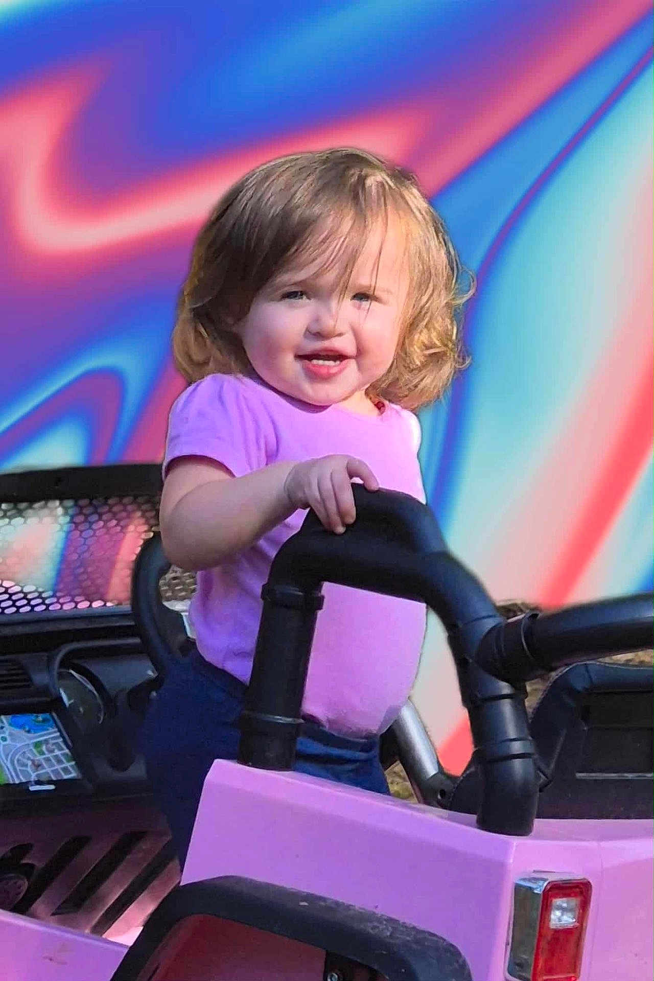 Madi joined the competition — help win amazing prizes! child, toddler, toy_car, purple_shirt, outdoor, smiling, play, vehicle, handle, happy, face, short_hair, daylight, colorful_background, plastic, black_handle, pink, person, cute, portrait