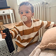 Gabriel a rejoint le concours — aidez-le/la à gagner de superbes lots ! toddler, pacifier, remote_control, striped_shirt, bed, blanket, pillow, curly_hair, carpet, table, chair, radiator, window, curtains, indoor, child, person, home, furniture, cozy
