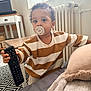 toddler, pacifier, remote_control, striped_shirt, bed, blanket, pillow, curly_hair, carpet, table, chair, radiator, window, curtains, indoor, child, person, home, furniture, cozy