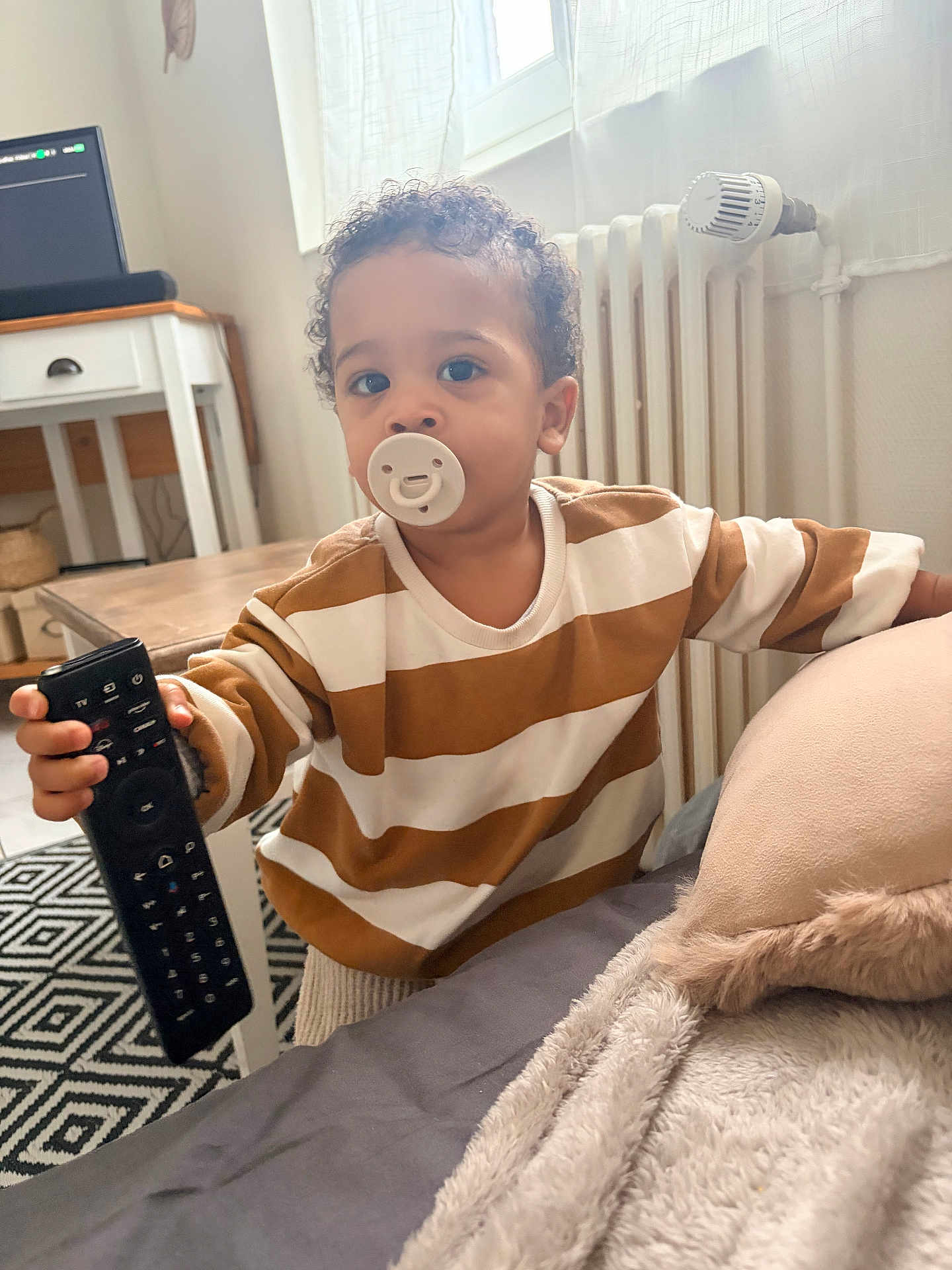 Gabriel a rejoint le concours — aidez-le/la à gagner de superbes lots ! toddler, pacifier, remote_control, striped_shirt, bed, blanket, pillow, curly_hair, carpet, table, chair, radiator, window, curtains, indoor, child, person, home, furniture, cozy