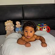 Damier is registered to the contest to win money with this photo: baby, child, curly_hair, pillow, plush_toy, stuffed_animal, mickey_mouse, lion_toy, bedroom, indoor, infant, cute, wide_eyes, portrait, furniture, soft_toys, couch, skin, expression, resting
