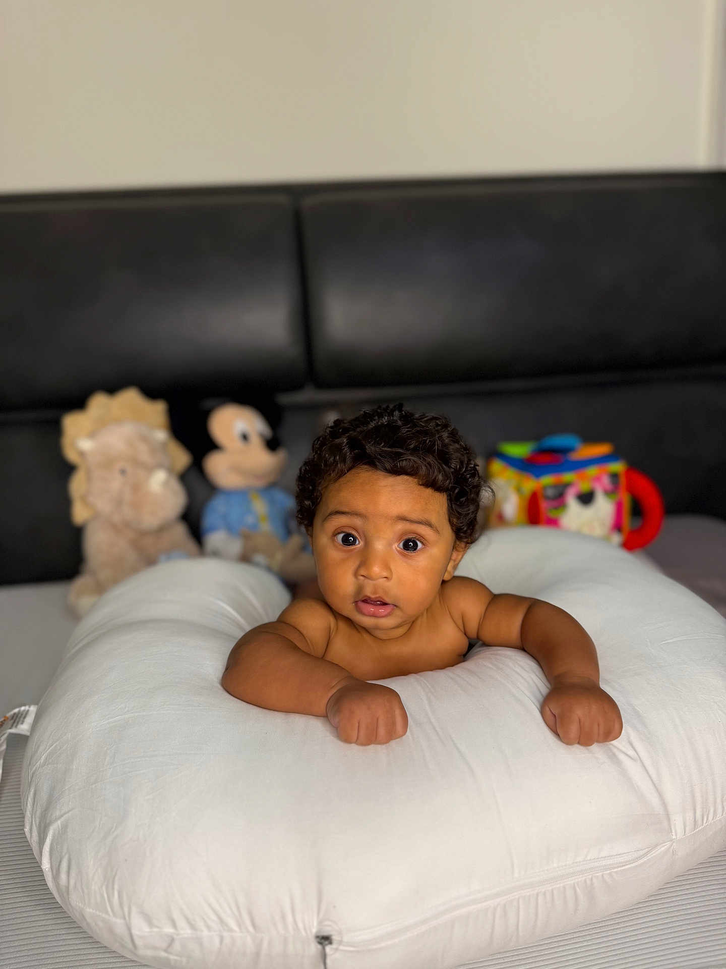 Damier is registered to the contest to win money with this photo: baby, child, curly_hair, pillow, plush_toy, stuffed_animal, mickey_mouse, lion_toy, bedroom, indoor, infant, cute, wide_eyes, portrait, furniture, soft_toys, couch, skin, expression, resting