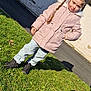 child, girl, braids, pink_coat, fur_hood, jeans, black_shoes, grass, shadow, sidewalk, outdoor, sunny, daylight, wall, curious_expression, person, casual_clothing, standing, nature, winter_clothing