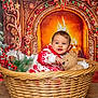 baby, basket, teddy_bear, christmas, fireplace, holiday_decorations, red_and_white_clothing, smiling, festive, pine_branches, gift_boxes, indoor, portrait, wooden_floor, cozy, toy, child, warm_lighting, decorative_ribbons, snowflake_ornaments