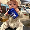 baby, child, infant, portrait, sitting, cap, hat, striped_outfit, clothing, blue_bucket, drinking, cup, store, workshop, indoor, industrial, face, hands, curious, funny