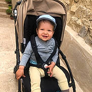 Léandre participe au concours pour gagner de l'argent avec cette photo : toddler, child, stroller, cap, smiling, happy, outdoor, stone_wall, potted_plants, pathway, clothing, shoes, baby, seatbelt, jacket, pants, footwear, daylight, person, urban