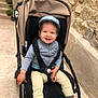 toddler, child, stroller, cap, smiling, happy, outdoor, stone_wall, potted_plants, pathway, clothing, shoes, baby, seatbelt, jacket, pants, footwear, daylight, person, urban