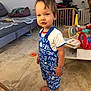 baby_gate, barefoot, blue_overall, casual, child, couch, face, floor, hair, home, indoor, living_room, person, pillow, playful, standing, table, toddler, toys, white_shirt