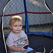 Colt is registered to the contest to win money with this photo: toddler, child, toy_car, trampoline, netting, outdoor, fence, light, shadow, casual_clothing, short_sleeves, pants, playtime, daylight, portrait, curious, sitting, young_child, blue_eyes, blond_hair