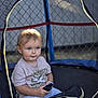 toddler, child, toy_car, trampoline, netting, outdoor, fence, light, shadow, casual_clothing, short_sleeves, pants, playtime, daylight, portrait, curious, sitting, young_child, blue_eyes, blond_hair