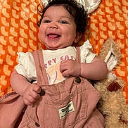 Alaya is registered to the contest to win money with this photo: baby, infant, smiling, curly_hair, bunny_ears, overalls, pink_clothing, white_shirt, plush_toy, stuffed_animal, orange_blanket, patterned_blanket, happy, cute, child, portrait, lying_down, indoors, soft_toy, toddler